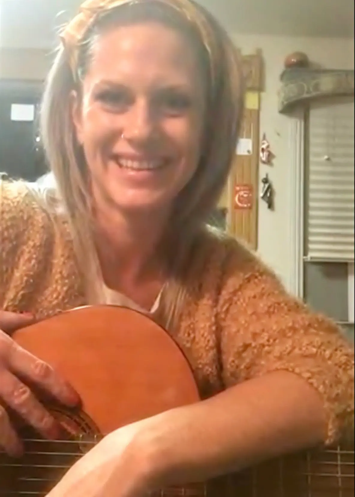 A smiling blonde-haired woman student holding a guitar during a fun, beginner-friendly online guitar lesson.