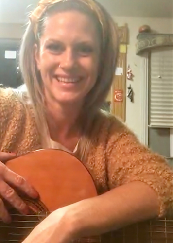 A smiling blonde-haired woman student holding a guitar during a fun, beginner-friendly online guitar lesson.